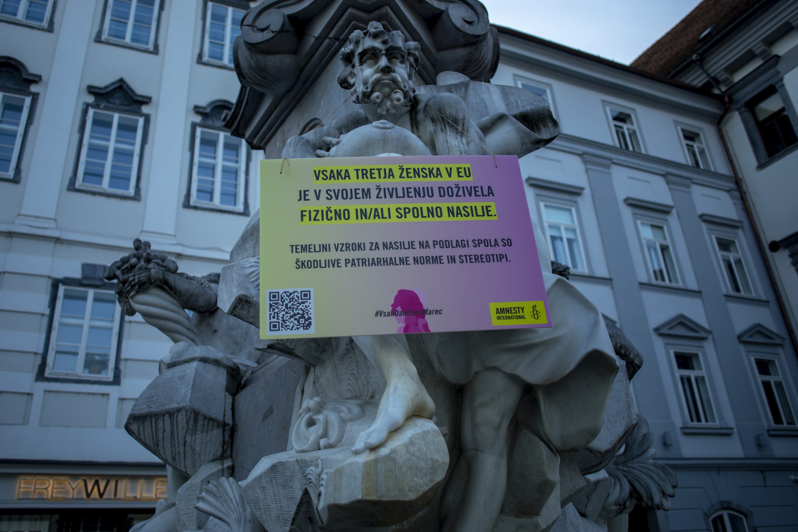 Pogled na Robbov vodnjak pred ljubljansko Mestno hišo, na katerem visi rumeno-roza tabla Amnesty Slovenije z napisom: "Vsaka tretja ženska v EU je v svojem življenju doživela fizično in/ali spolno nasilje. Temeljni vzroki za nasilje na podlagi spola so škodljive patriarhalne norme in stereotipi. #vsakdanosmimarec"