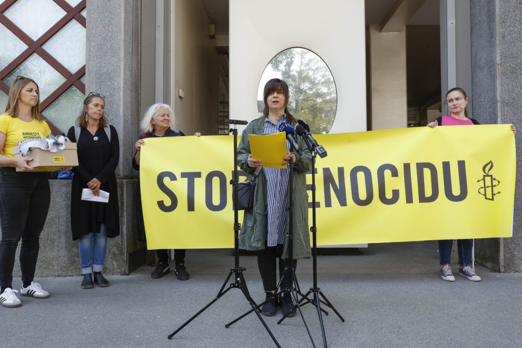Skupina ljudi v ozadju drži napis STOP GENOCIDU in škatlo s podpisi peticije, na katero je simbolno položena palestinska kefija. Direktorica AIS pred mikrofoni vlado RS poziva k ukrepanju.