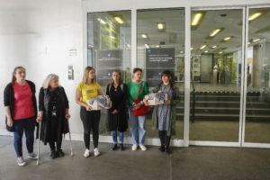 Skupina ljudi stoji pred vhodom vlade RS. V rokah držijo dve škatli s podpisi, na kateri sta simbolno položeni palestinski kefiji.