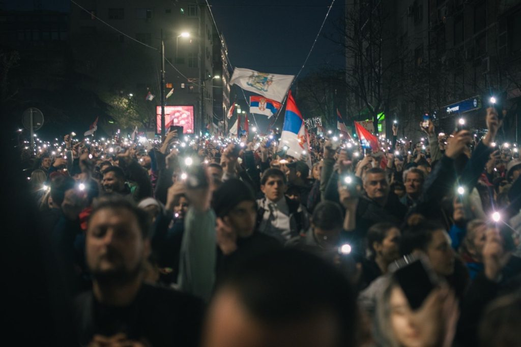 Množica ljudi po temi protestira. V rokah dvigajo telefone in svetijo z lučkami in vihtijo srbske zastave.