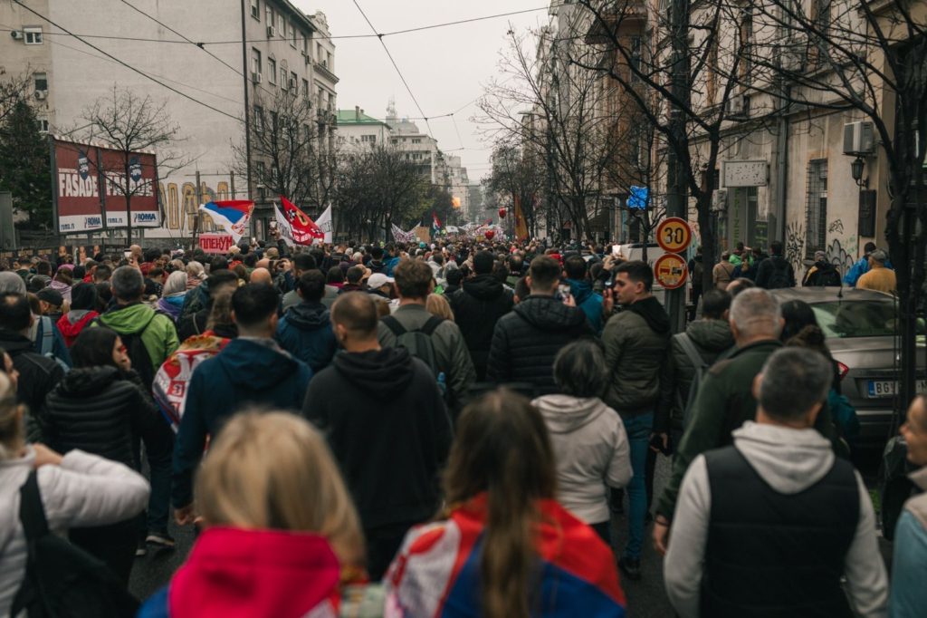 Pogled na množico ljudi v hrbet, ki po dnevu mirno protestira. Ozadje je mestno jedro. V zraku plapola več zastav.