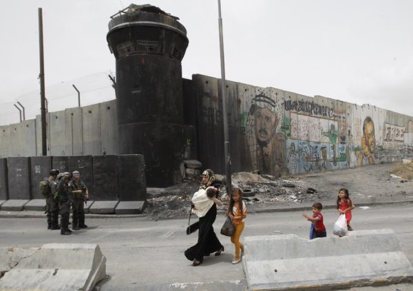 Palestinka in otroci hodijo mimo pripadnikov izraelskih varnostnih sil, ki stojijo na straži pred kontroverzno izraelsko ločilno ograjo na kontrolni točki Qalandia blizu mesta Ramala na Zahodnem bregu 5. junija 2014.