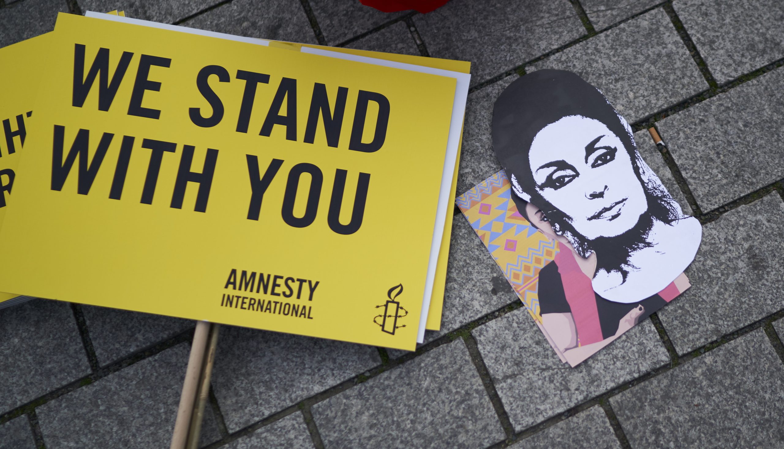 Rumen transparent z napisom 'WE STAND WITH YOU' in slika Eren Keskin.