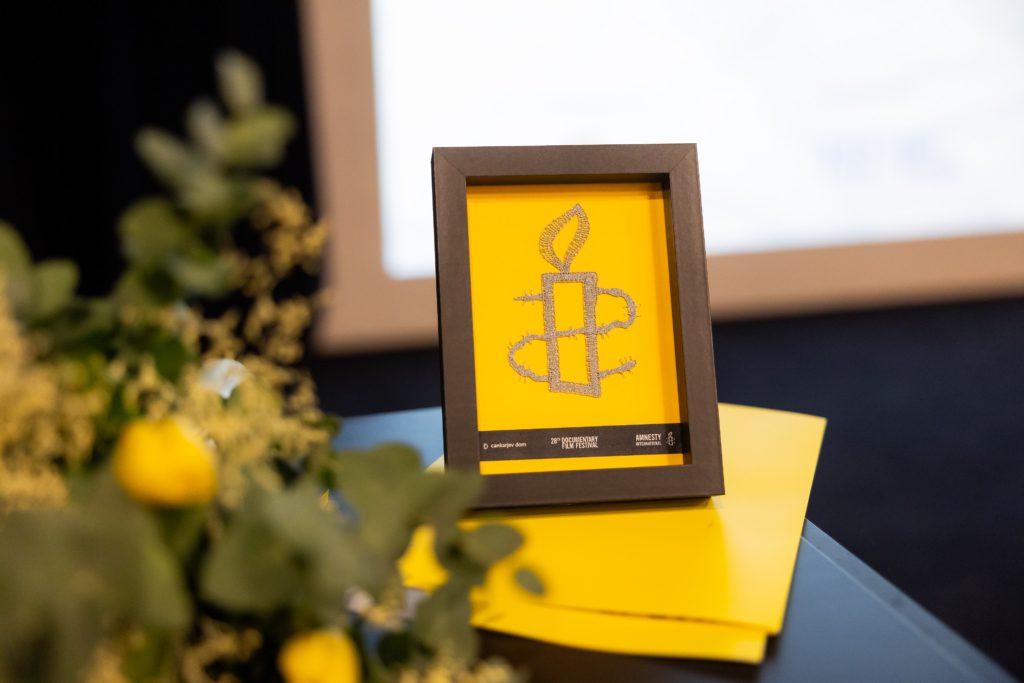 Srebrna čipka v obliki svečke na rumeni podlagi na mizi, poleg šopek. Nagrada Amnesty International Slovenije na Festivalu dokumentarnega filma.