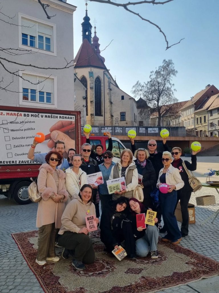 Skupina nasmejanih ljudi pred kombijem s plakati, ki pozivajo k udeležbi na volitve.