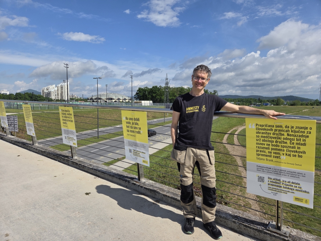 Prostovoljec stoji pred osveščevalnimi tablami, v ozadju silhueta mesta.